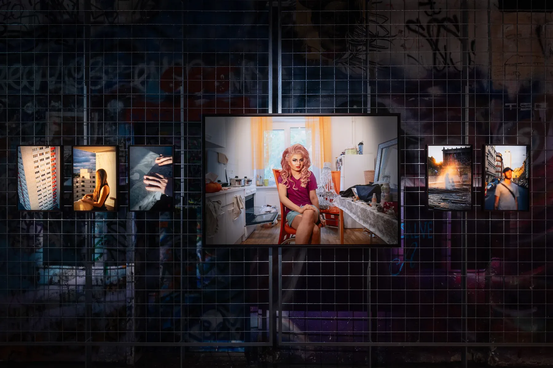 Installation view: a row of prints on a dark mesh screen, centred on a large portrait of a figure seated in a sunlit kitchen.