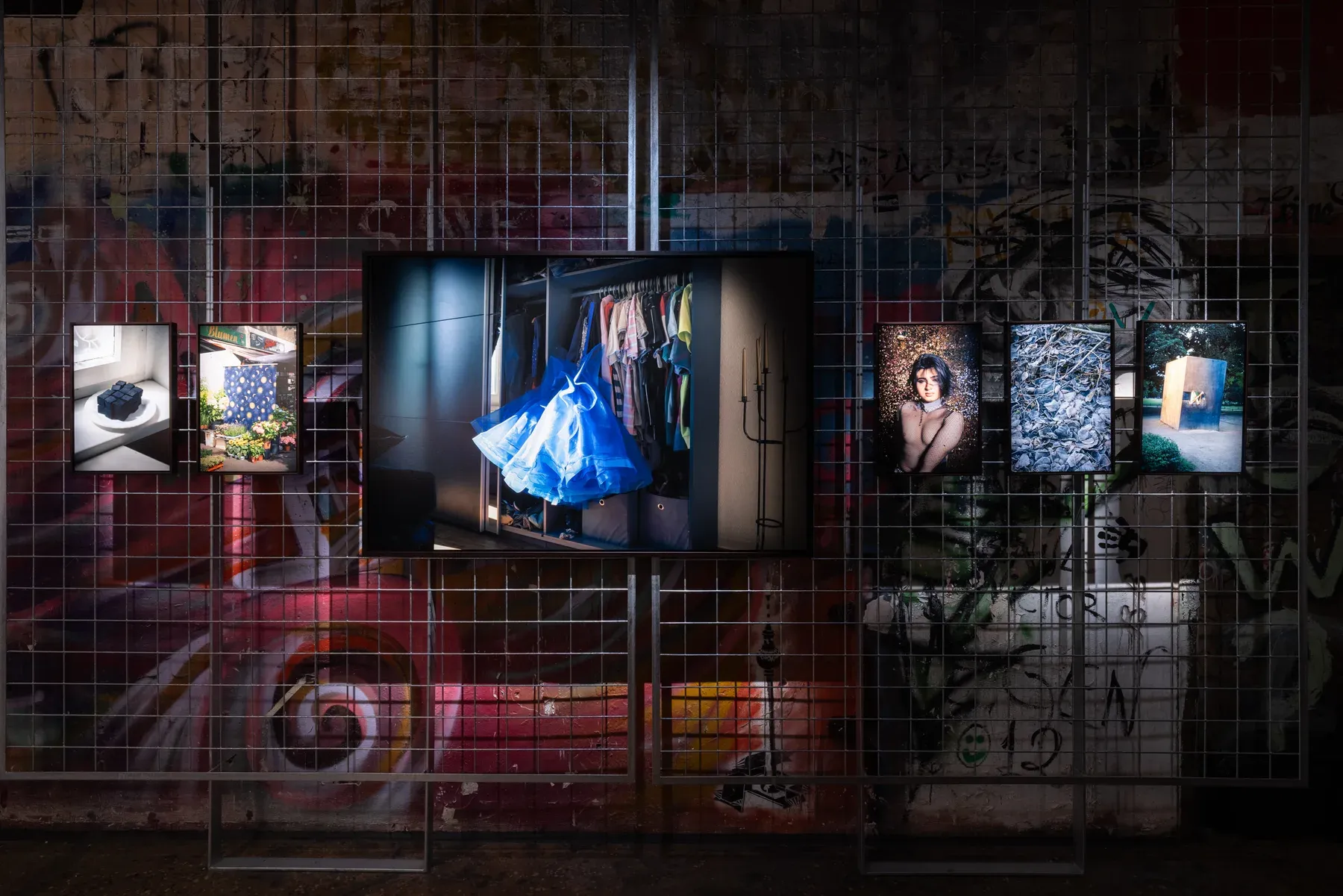 Installation view: prints hung on a steel mesh screen against a red graffiti mural, centred on a large photograph of a blue dress caught mid-swing in an open wardrobe.