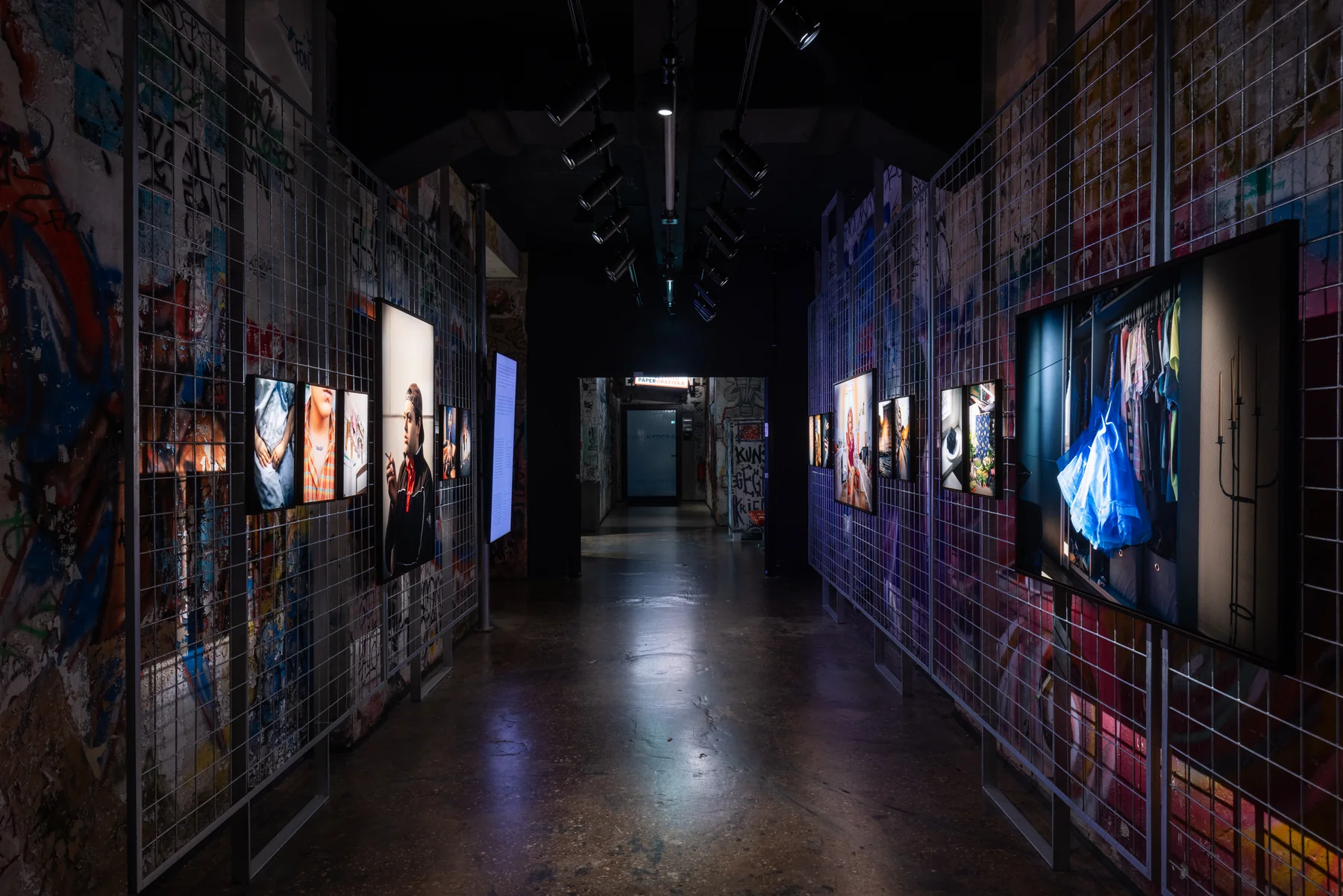 Installation view: the corridor of the exhibition, lit prints running down both graffiti-covered walls.