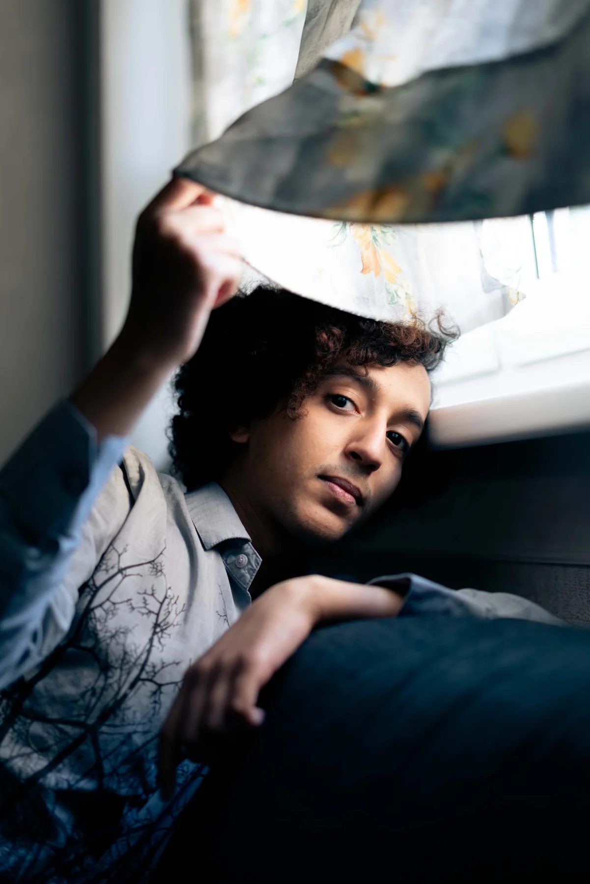 A person reclines by a window, holding a patterned garment above their head.
