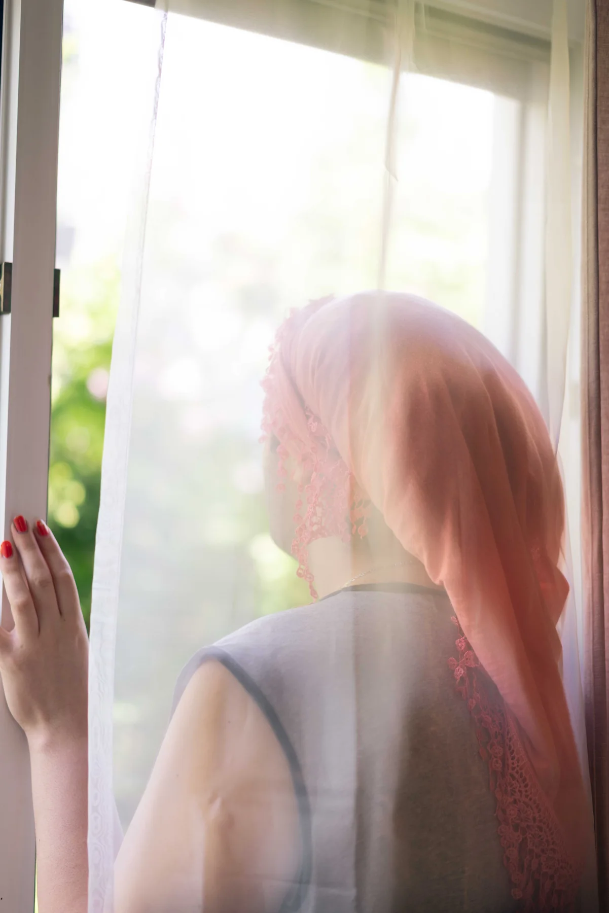A person in a pink headscarf looks out through a curtained window.