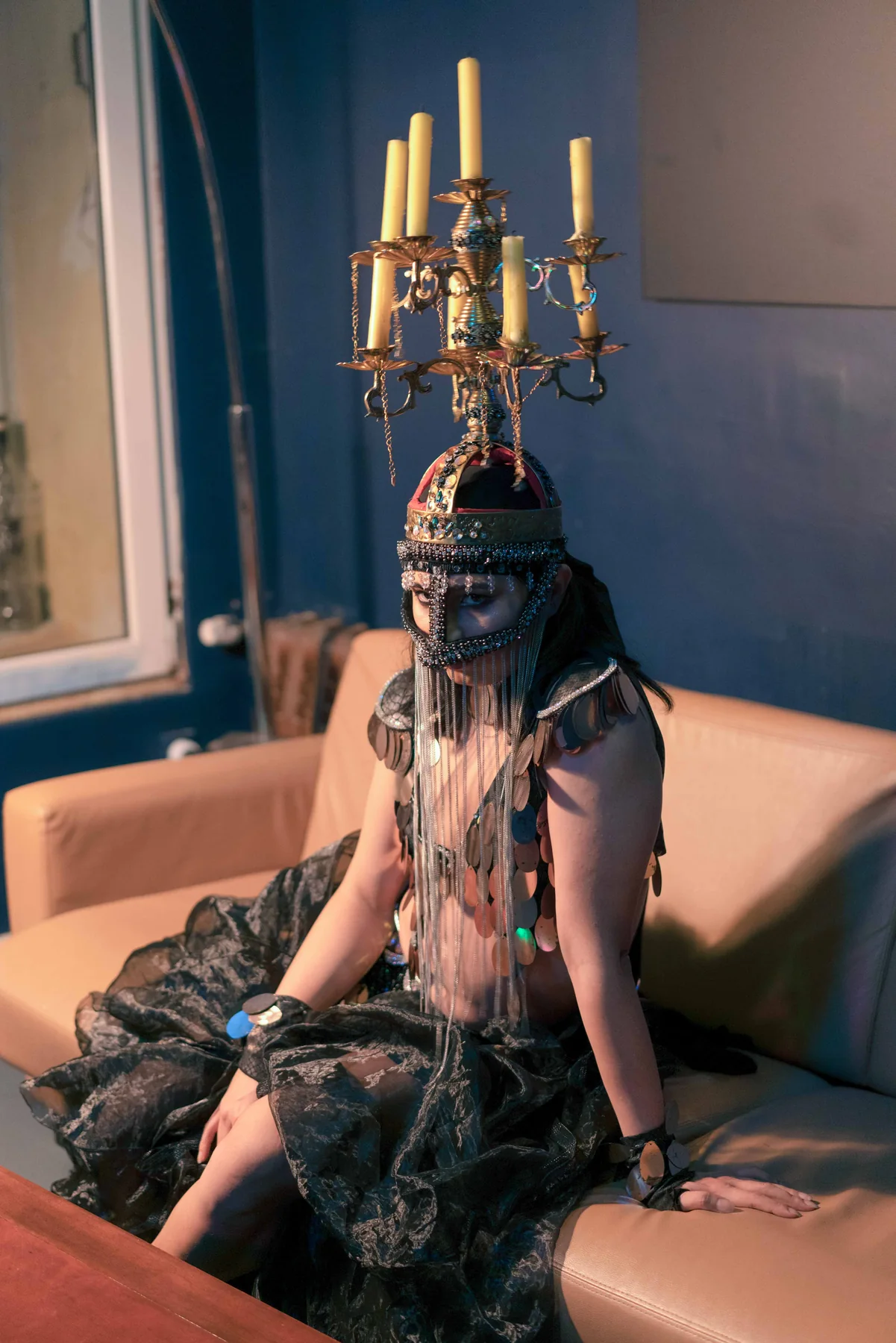 A person in costume sits on a sofa beneath a candelabra headdress.