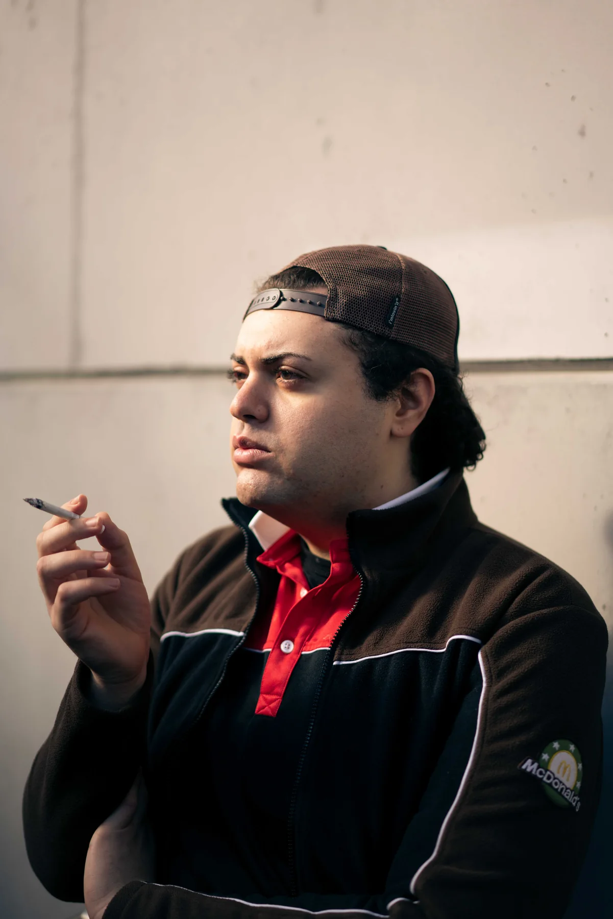A person in a McDonald's work jacket and backwards cap smokes against a concrete wall.