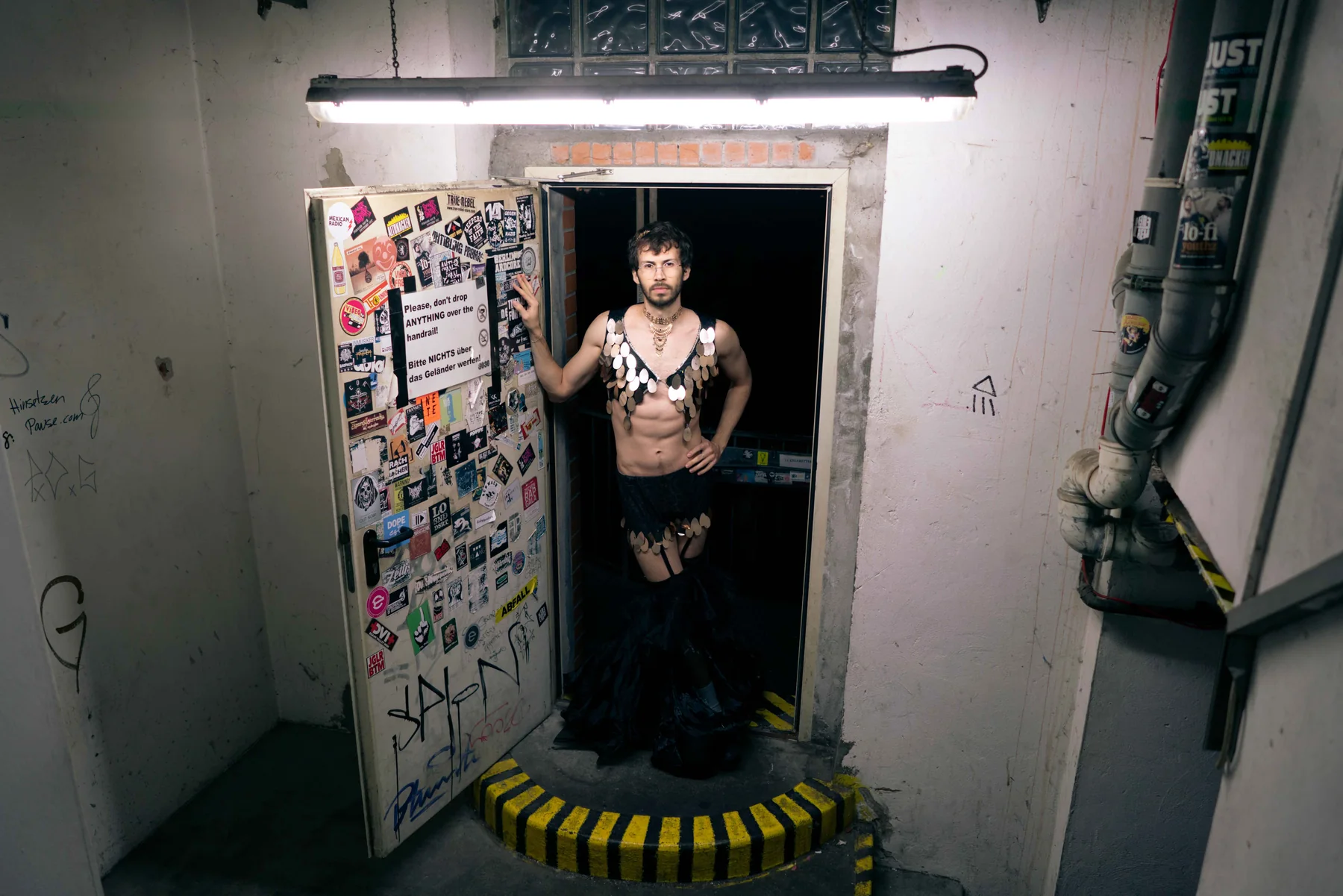 A person stands in a lit basement doorway covered in stickers, wearing a beaded fringe costume.