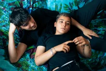 Two people in black t-shirts lie on a turquoise floral bedspread, fingertips touching, one looking at the other.