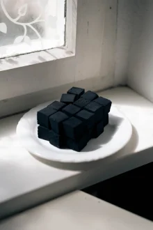 Black charcoal cubes stacked on a white plate on a sunlit windowsill beside a frosted window.