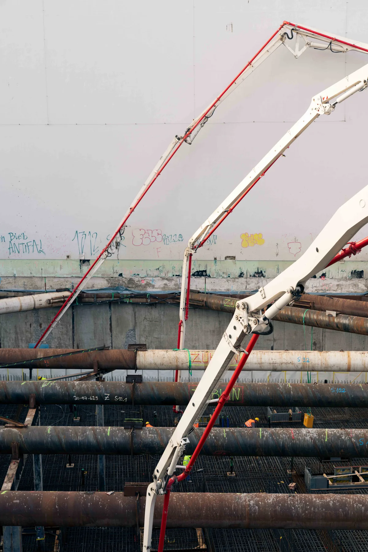 A red and white concrete pump arm crosses a construction pit spanned by rusted steel pipes, workers in high-visibility vests small below.