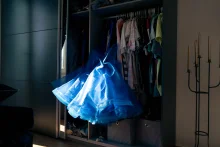 A blue tulle dress hangs in a patch of light at an open wardrobe of clothes.