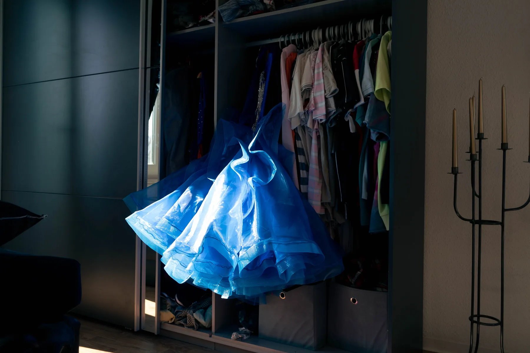 A blue tulle dress hangs in a patch of light at an open wardrobe of clothes.