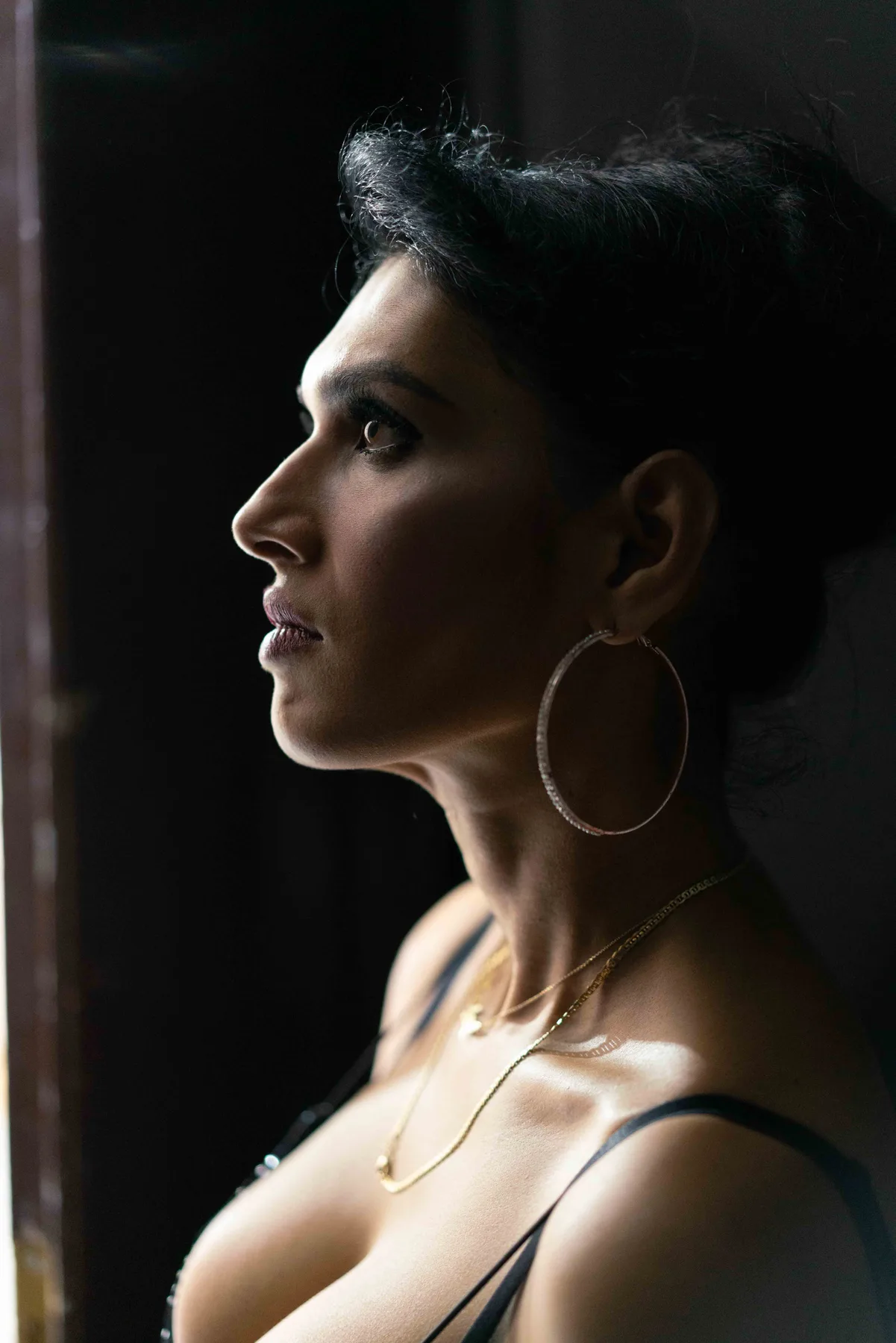 A person in profile in a dark room, hair swept back, wearing a large hoop earring and gold chains.