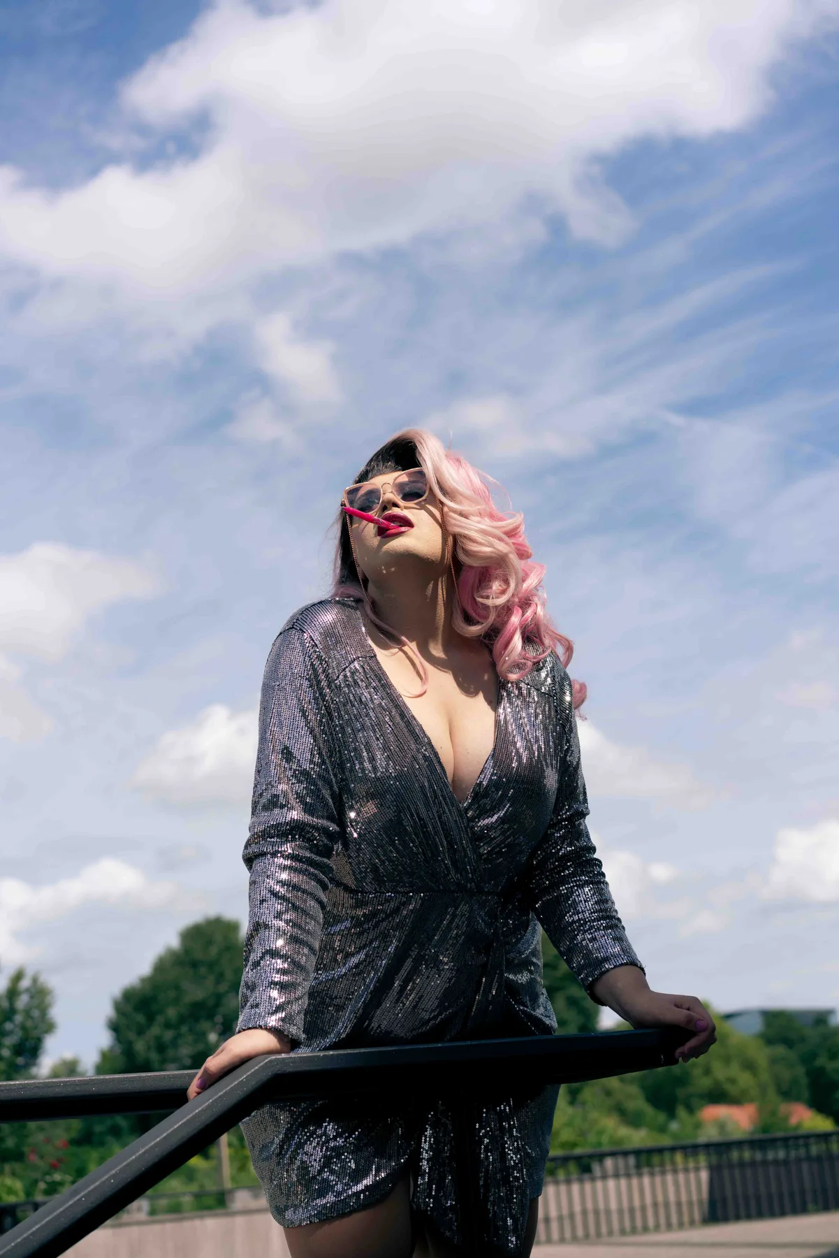 A person in a silver sequinned dress, pink wig and sunglasses leans back against a railing under a bright sky.