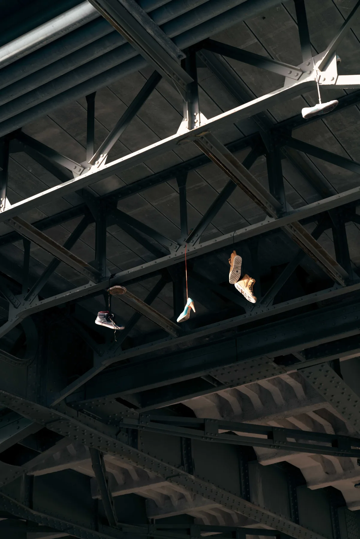 Trainers and a high-heeled shoe hang by their laces from the sunlit steel girders under a bridge.