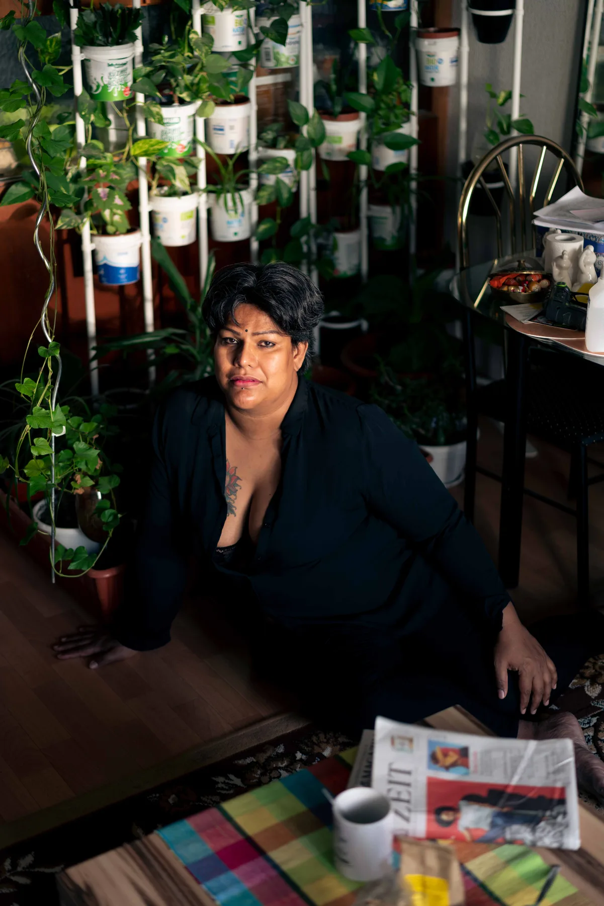 A person in an open black shirt sits on a wooden floor beside a wall of potted plants in yoghurt tubs.