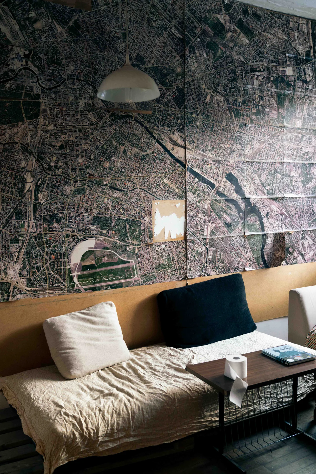 A pendant lamp hangs over a low bed with cushions in a room papered with aerial city maps.