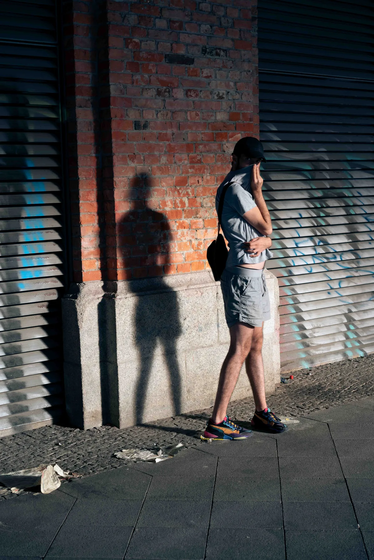 A person in a black cap holds their grey T-shirt to their face, shadow falling on the sunlit brick wall.