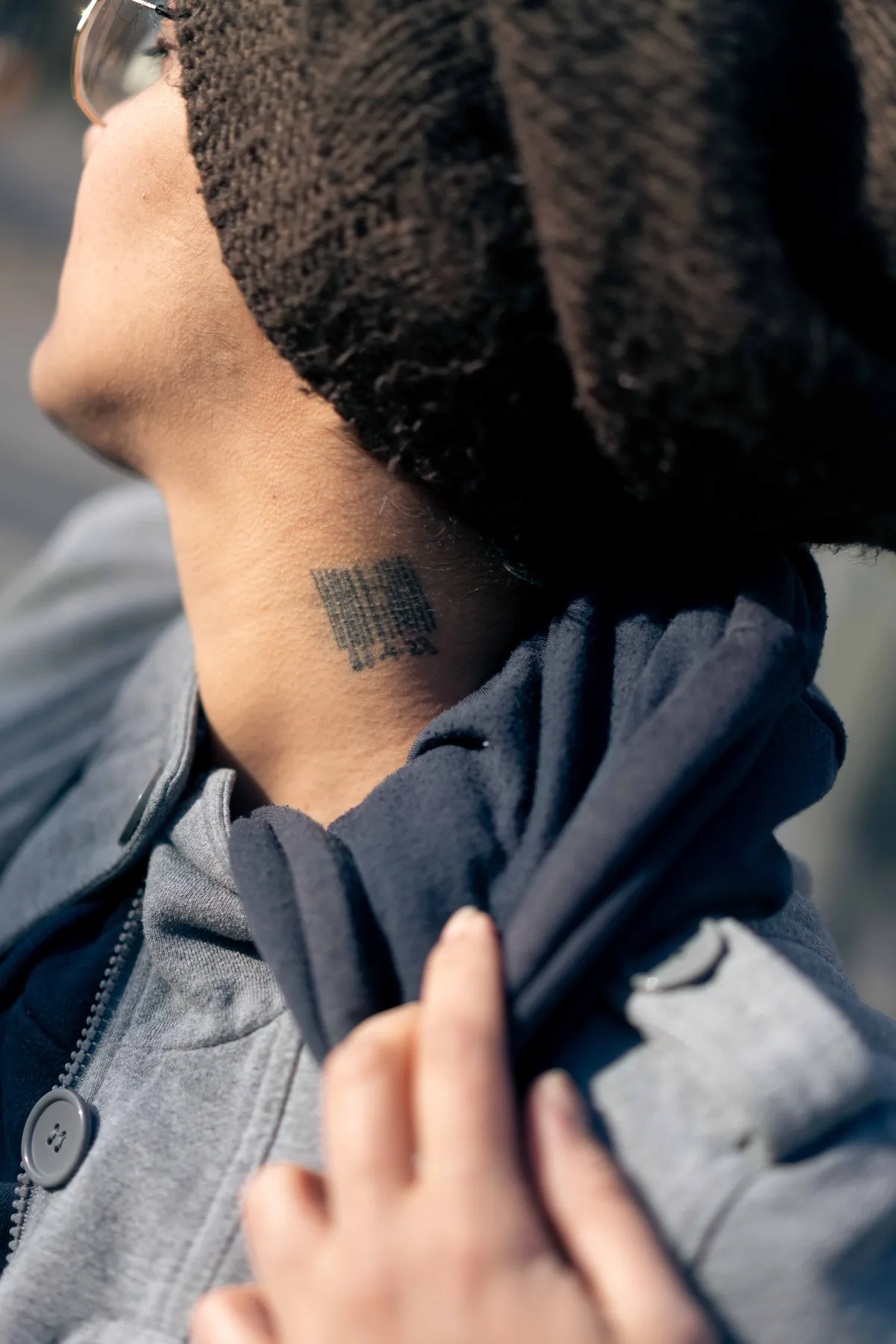 A person's neck in bright sunlight, a small block tattoo below dark curls, one hand at their collar.