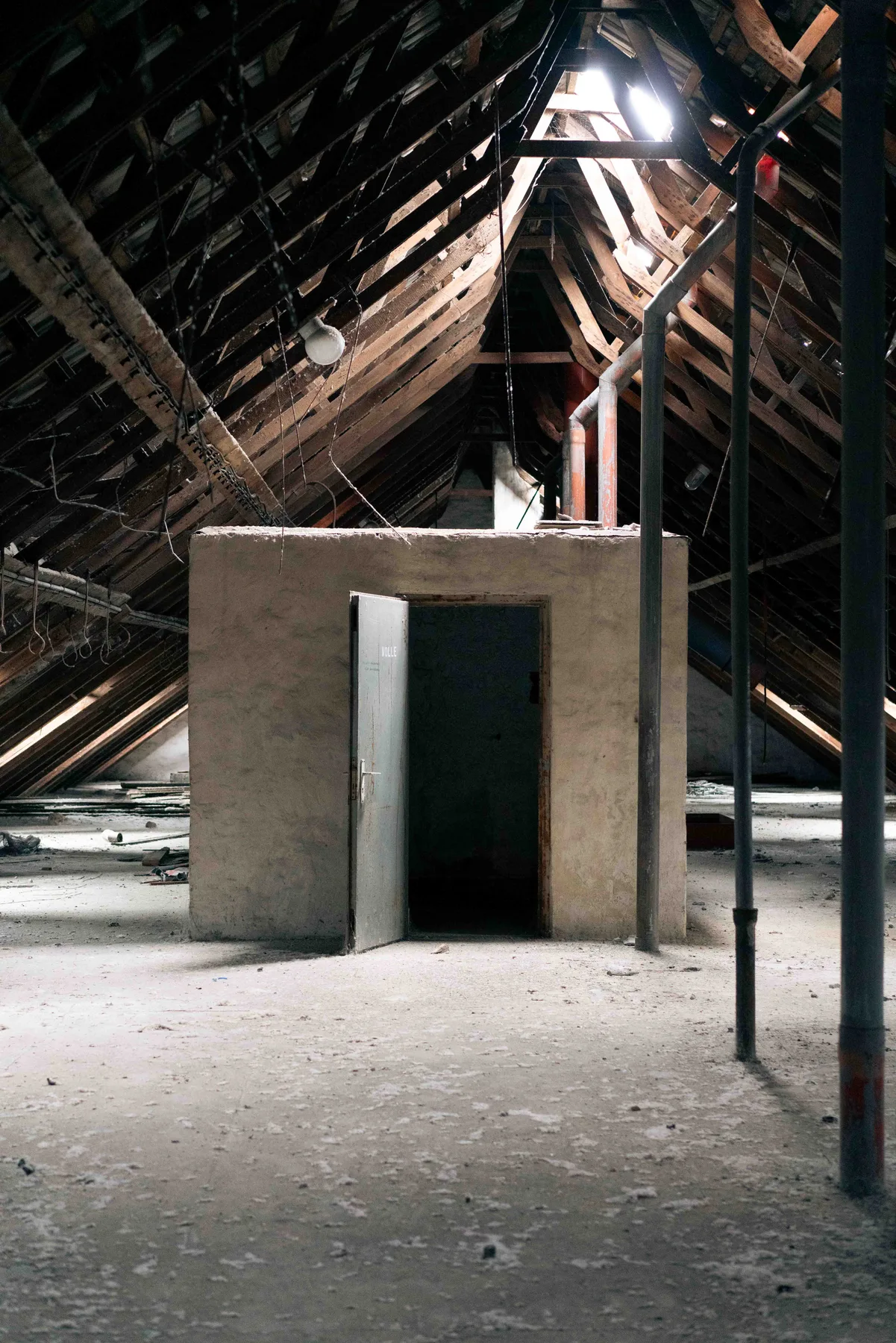 A metal door stands ajar in a plastered enclosure under the bare rafters of a dusty attic.