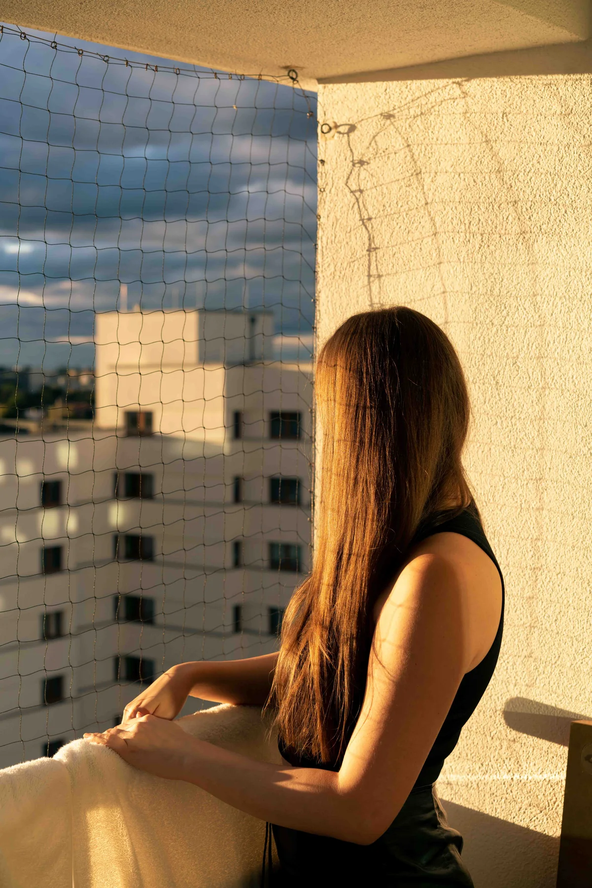 A person with long hair leans on a netted balcony rail in low sun, facing an apartment block.