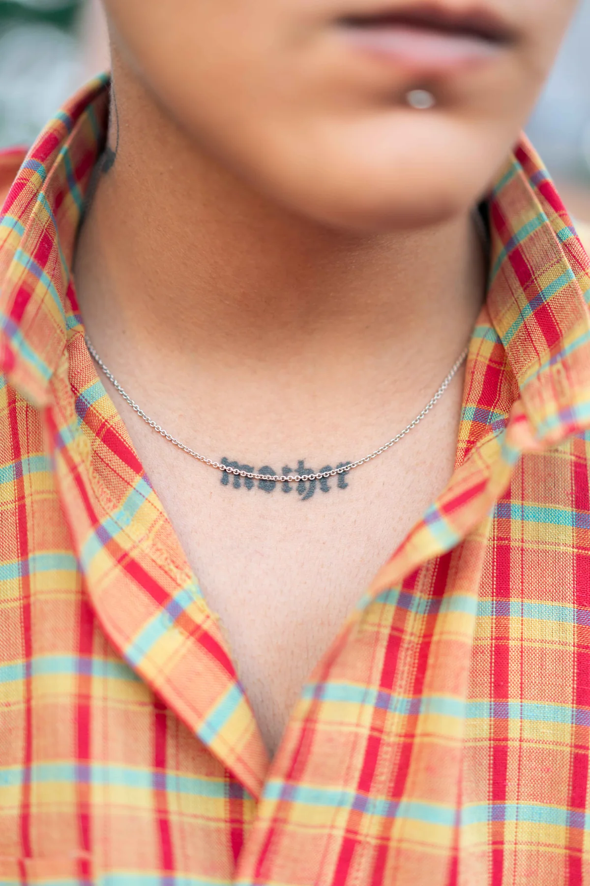 A tattoo reading mother at the base of a neck, under a silver chain and open plaid collar.