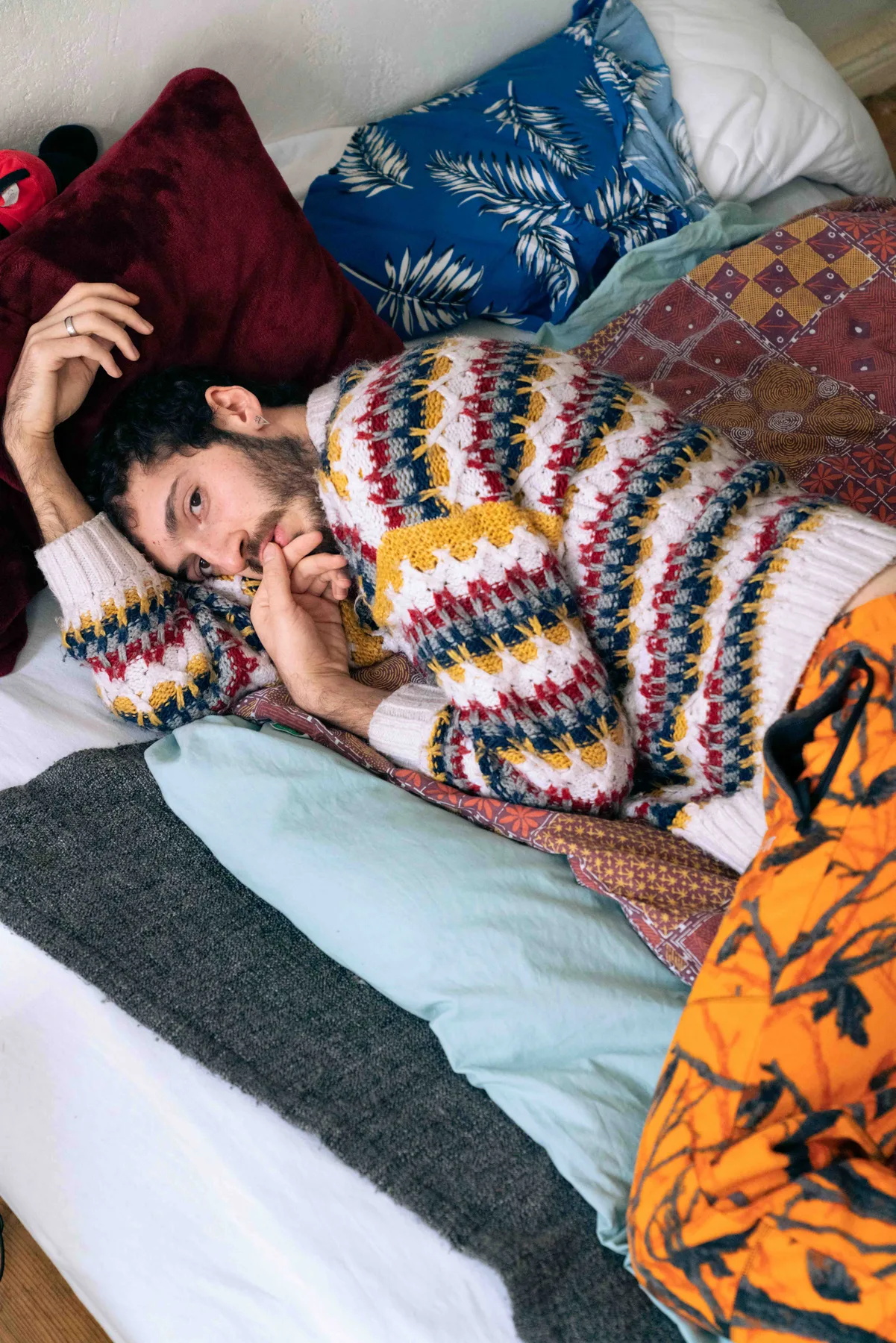 A person in a patterned knit sweater lies on a mattress among pillows, one hand at their mouth.
