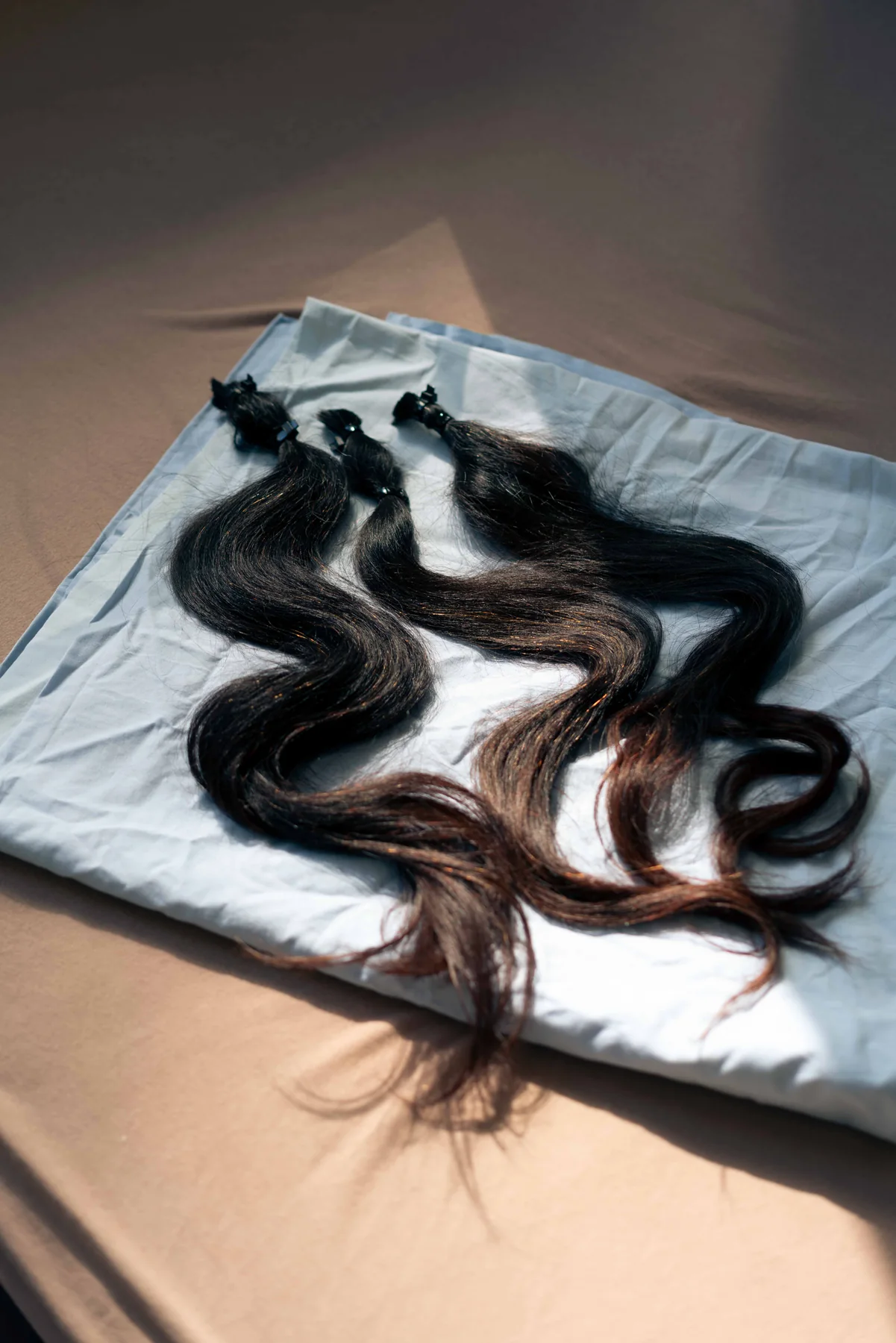 Three lengths of dark wavy hair extensions, clipped at one end, lie on a sunlit white sheet.