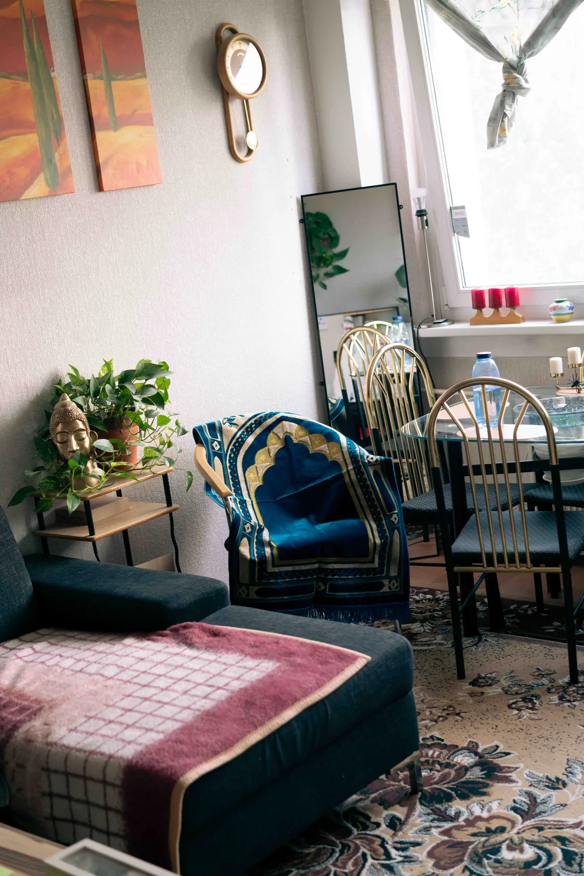 A blue and gold embroidered rug is draped over a chair beside a dark sofa and a bright window.