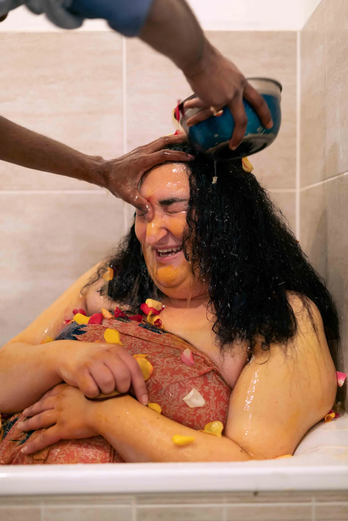 Hands pour water from a blue bowl over a person in a bathtub, yellow paste and petals on their skin.