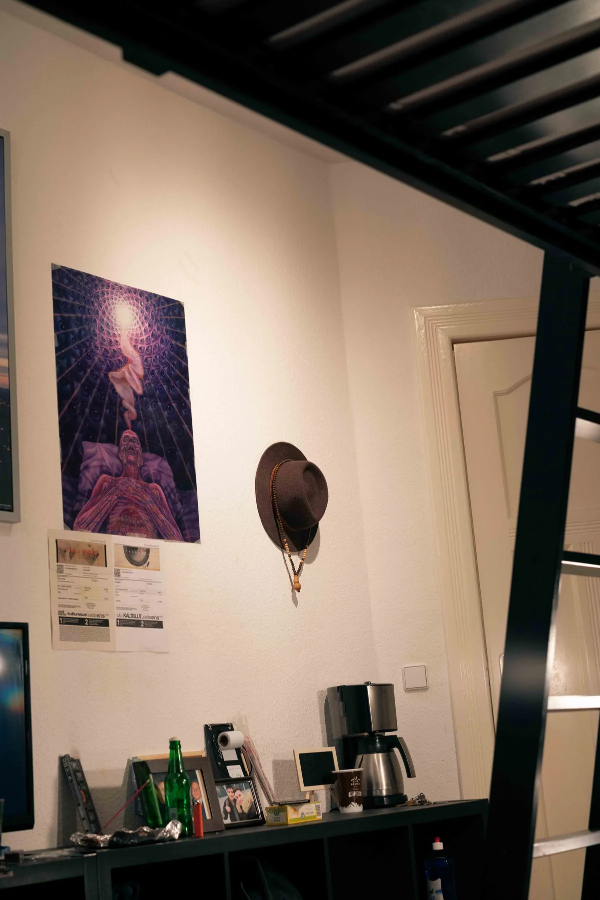 A brown felt hat and beaded necklace hang on a white wall above framed photos and a coffee maker.
