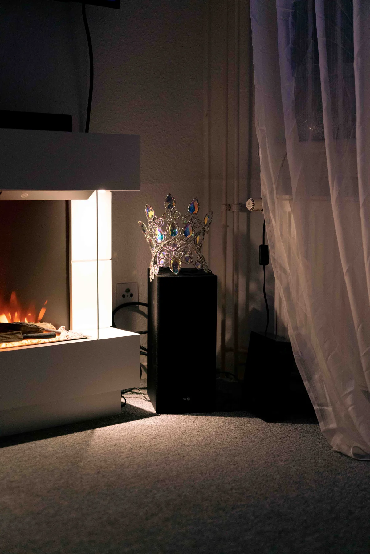 A rhinestone crown sits on a black speaker beside a lit electric fireplace, sheer curtains to the right.