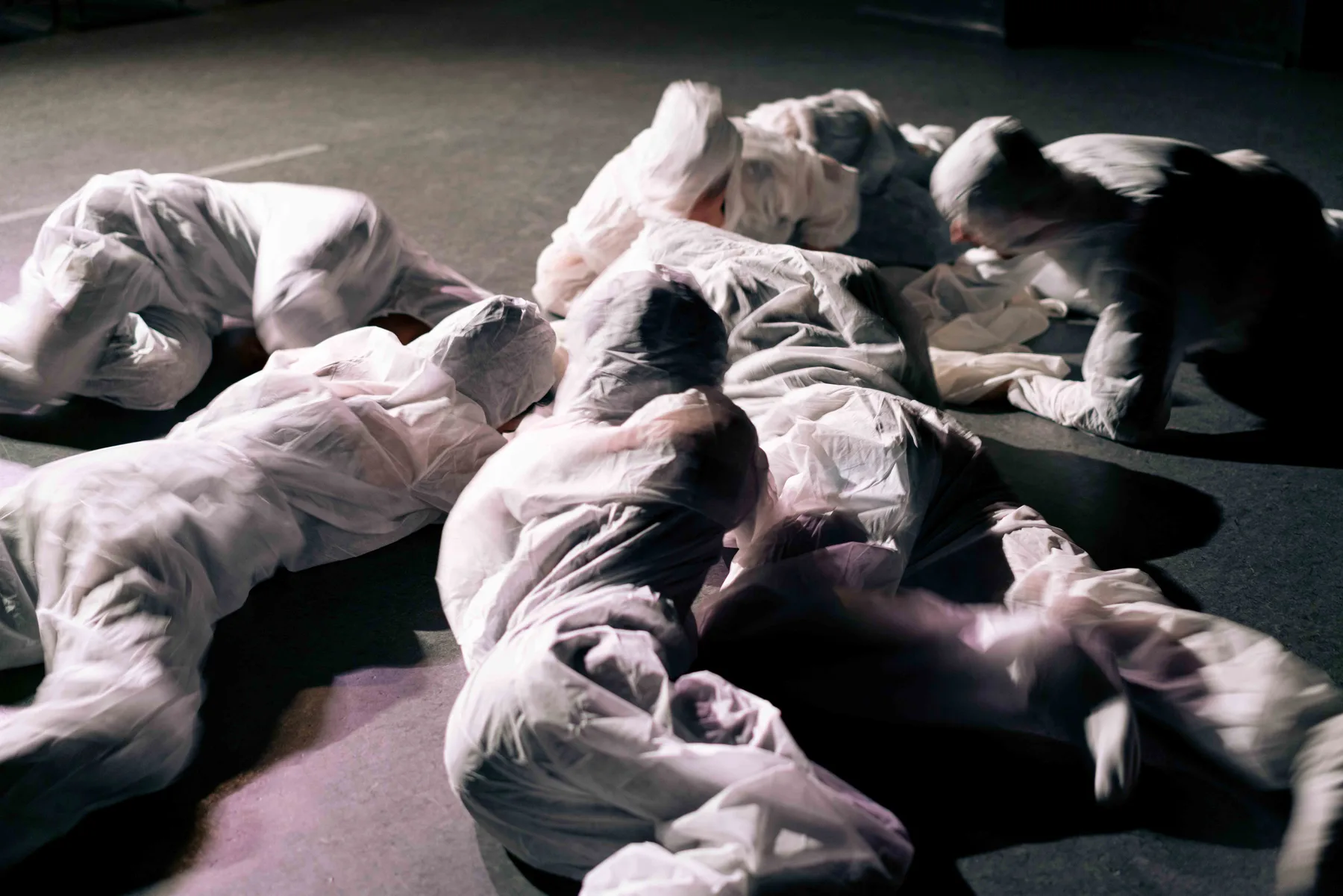 Several people wrapped in white sheets lie and crouch on a dark floor, blurred by movement.