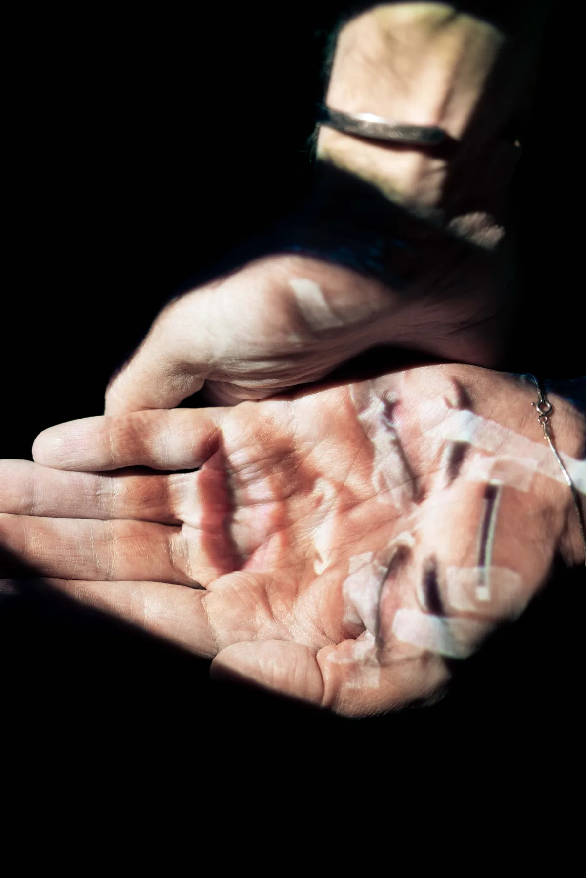 Two open palms held out in darkness, light falling in patches across the fingers and a silver bracelet.