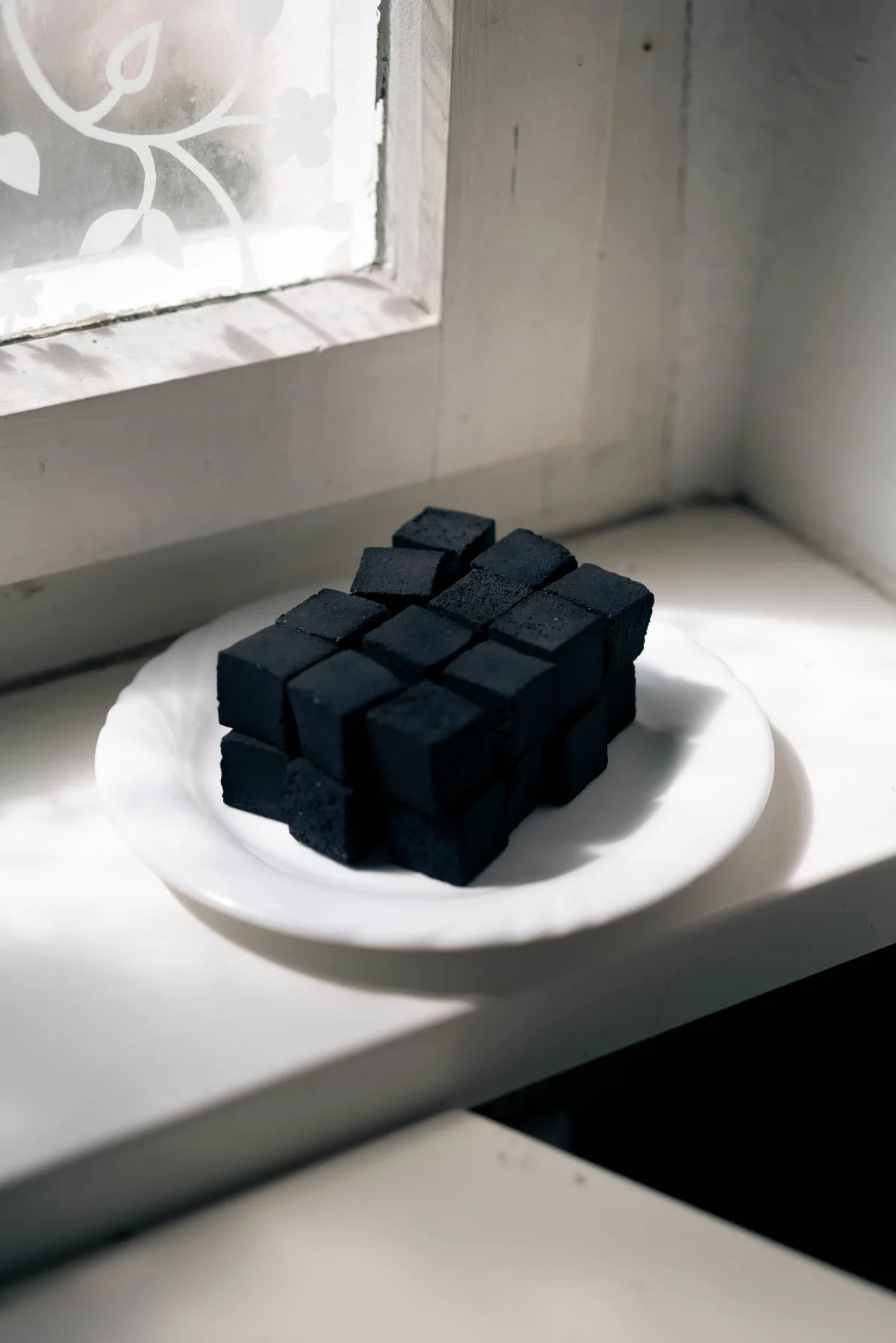 Black charcoal cubes stacked on a white plate on a sunlit windowsill beside a frosted window.