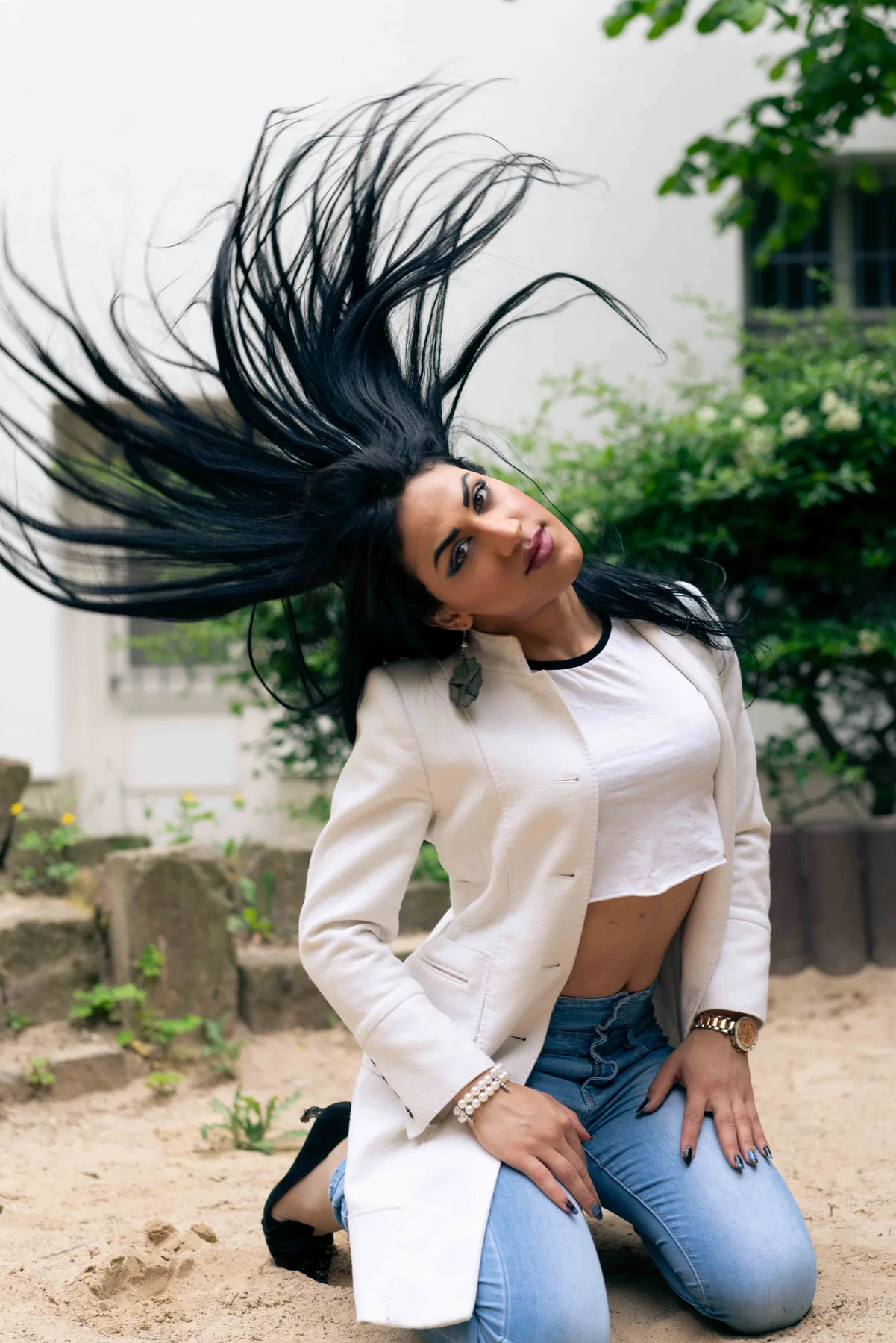 A person in a white blazer and jeans kneels on sand, long black hair thrown up in the air.