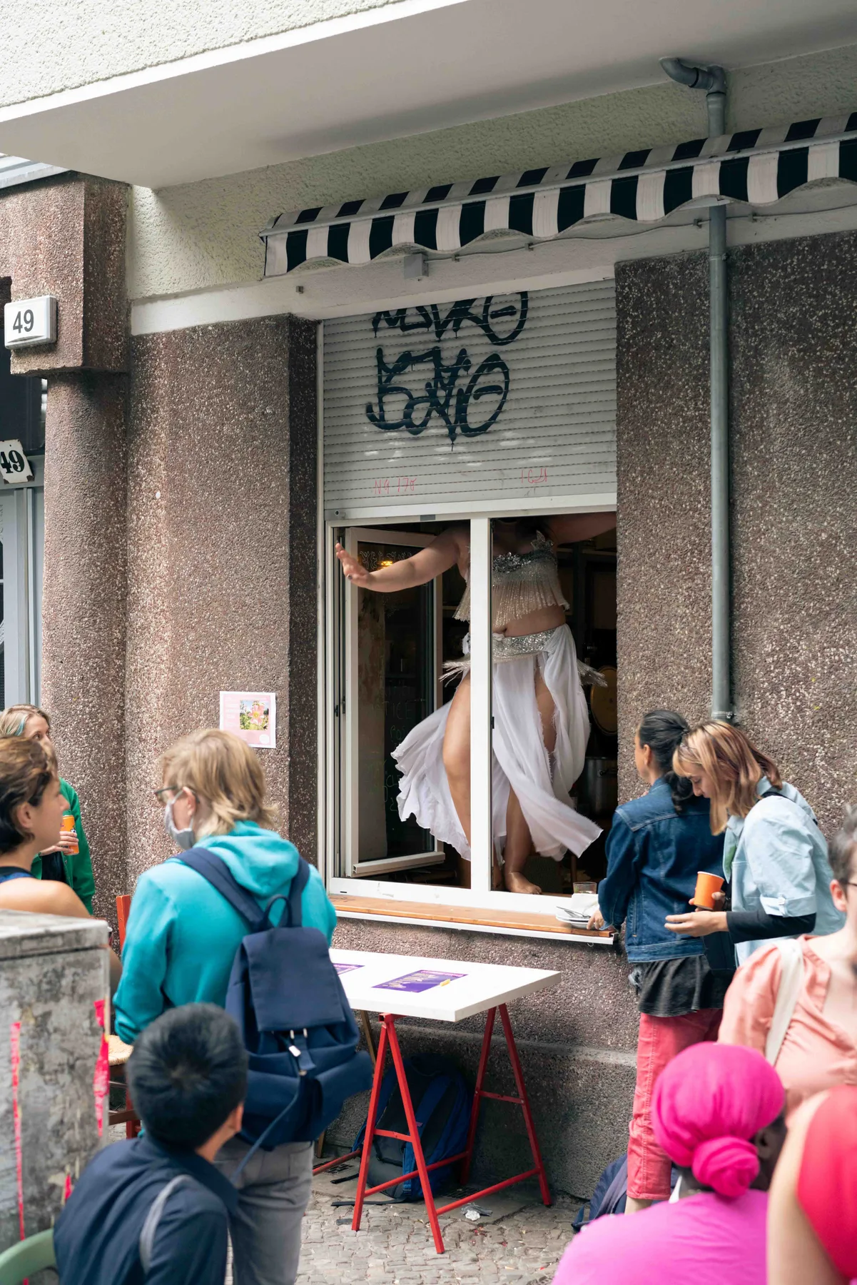 A person in a silver beaded top and white skirt stands barefoot in an open window above a crowd.