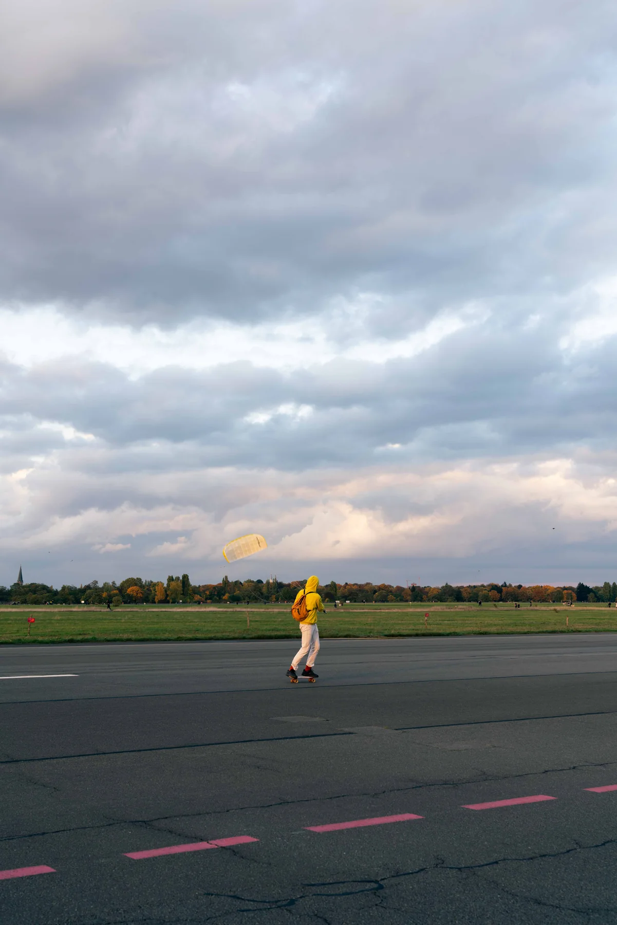 A person in a yellow hooded jacket skates along an empty runway, flying a yellow kite under thick cloud.