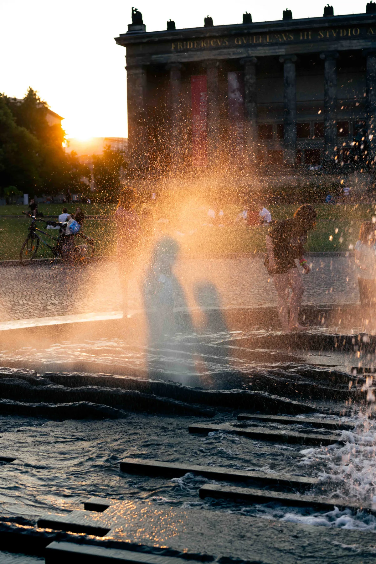 A person stands in the spray of a fountain at sunset, a colonnaded museum building behind them.