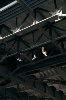 Trainers and a high-heeled shoe hang by their laces from the sunlit steel girders under a bridge.