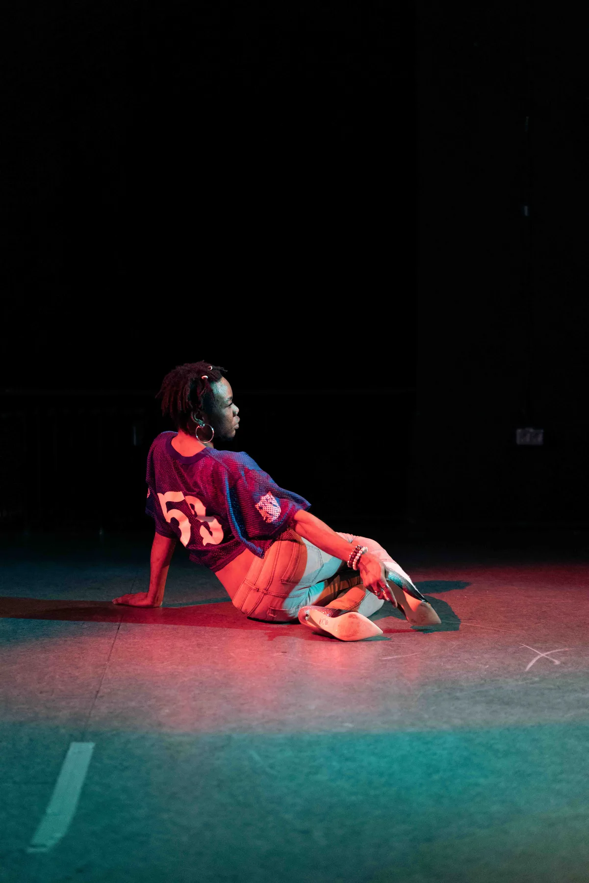 A person in a mesh jersey sits on a stage floor lit red and teal, reaching for a silver heel.