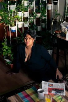 A person in an open black shirt sits on a wooden floor beside a wall of potted plants in yoghurt tubs.