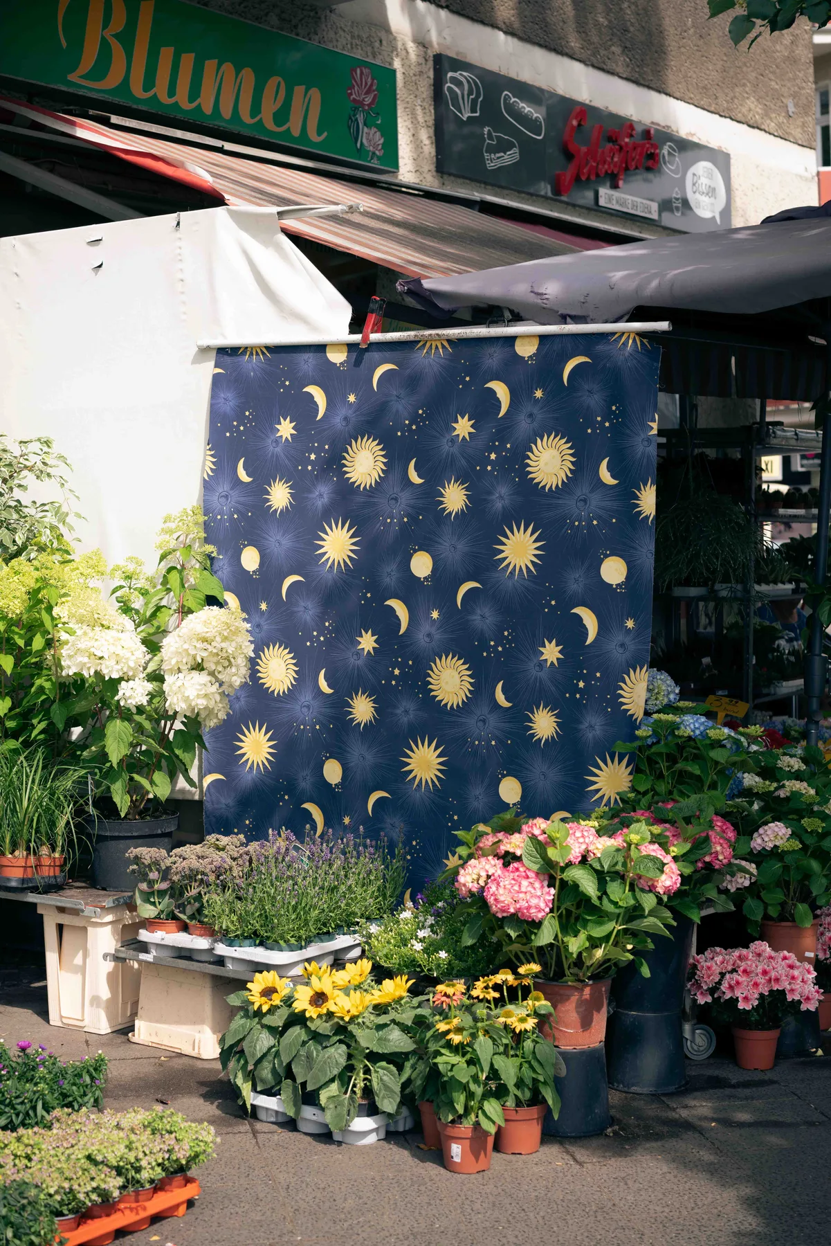 A navy cloth printed with gold suns, moons and stars hangs at a flower stall among potted hydrangeas and sunflowers.