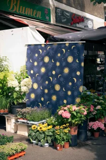 A navy cloth printed with gold suns, moons and stars hangs at a flower stall among potted hydrangeas and sunflowers.