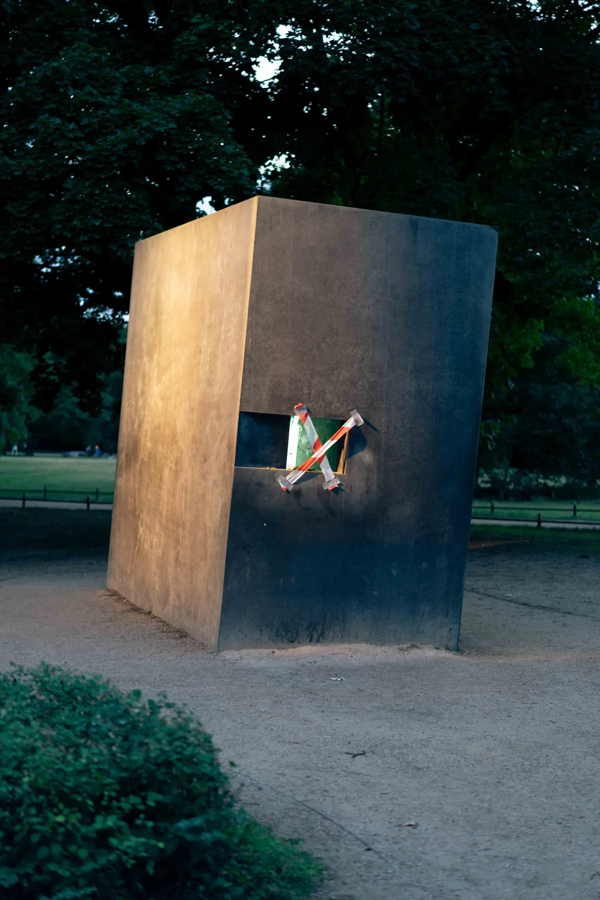 A dark metal cube stands on gravel in a park, its small window crossed with red-and-white tape.